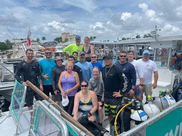 Group of people posing on a boat with scuba tanks and gear, under a partly cloudy sky.