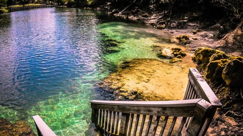 Wooden stairs leading into a clear, blue-green natural spring surrounded by rocks and shaded trees.