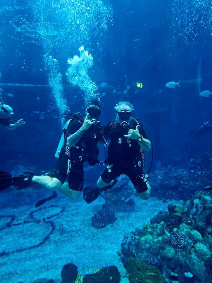 Two scuba divers underwater giving hand signals near coral reef, surrounded by bubbles.