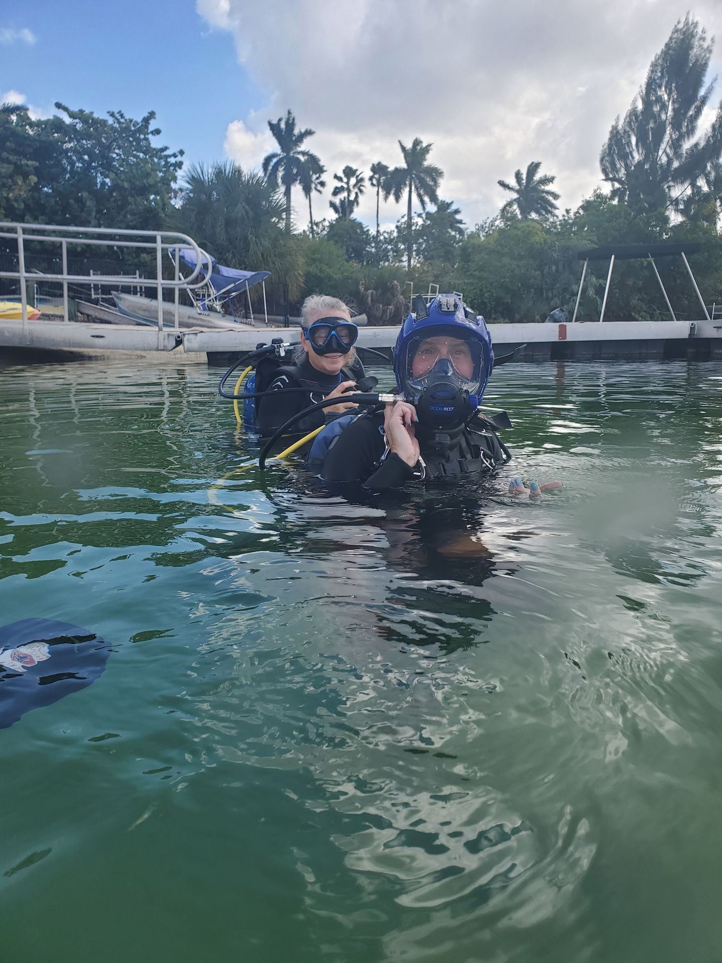 Two scuba divers in water near a dock, surrounded by tropical trees and a cloudy sky.