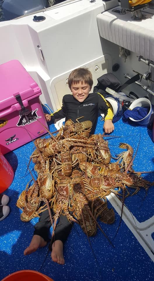 Child lying on boat floor covered with numerous lobsters, next to pink cooler in bright sunlight.