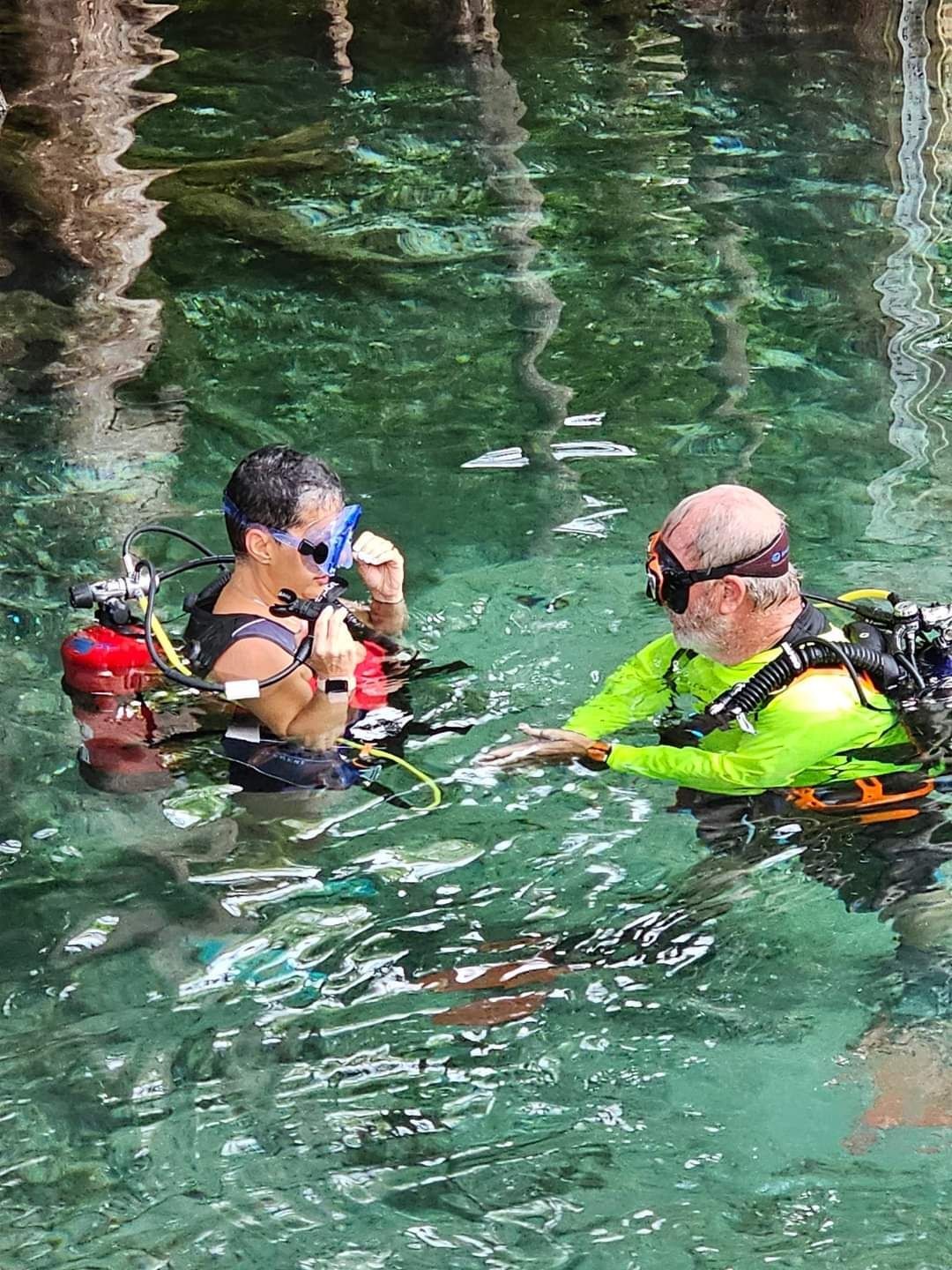 Two scuba divers in clear water, one adjusting mask, the other assisting.