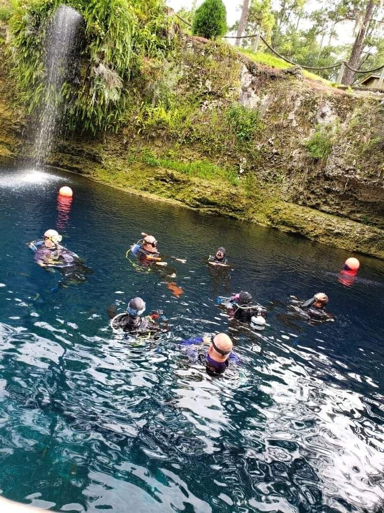 Group of scuba divers in open water near a small waterfall and rocky ledge with greenery.