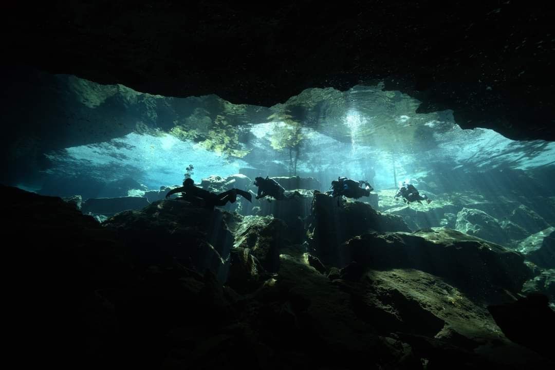 Group of scuba divers exploring an underwater cave with beams of sunlight penetrating the clear blue water.